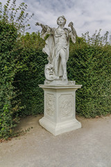 Female Statue in beautiful garden. Monument made of white stone stands on rectangular pedestal. Statue of woman which holds out one of her hand and the othe hand holds a shell
