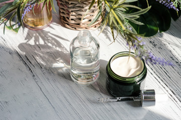 Cosmetic cream container and a bootle of serum with green herbal leaves on white table