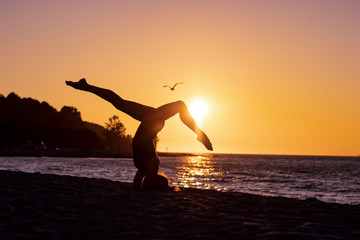 Fototapeta premium Woman doing gymnastic yoga on the beach at sunset, orange sky in the background with a bird flying in shadow