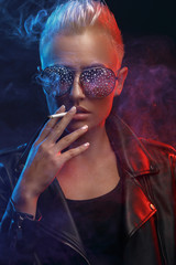 Portrait of a smoking blonde girl with stylish sunglasses on a dark background. Creative color. Studio photography.