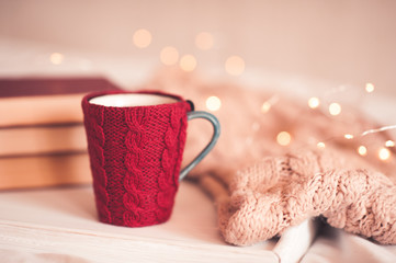 Obraz na płótnie Canvas Red knitted cup of coffee with stack of book and knitted scarf over Christmas lights close up. Winter holiday season. Good morning. Glowing Christmas garland.