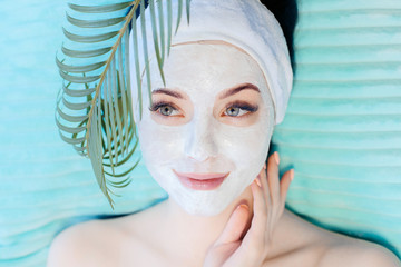 Portrait of happy beautiful woman holds green tropical leaf. Young girl enjoys cream facial mask. Natural cosmetics, skincare, wellness, beauty clinic, facial treatment, cosmetology concept.