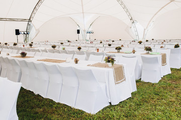 View over long table with white tablecloth covered with brown napkins and flower pots on the grass under the roof.