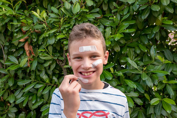 Boy with plaster on his face in front of a hedge looks sad