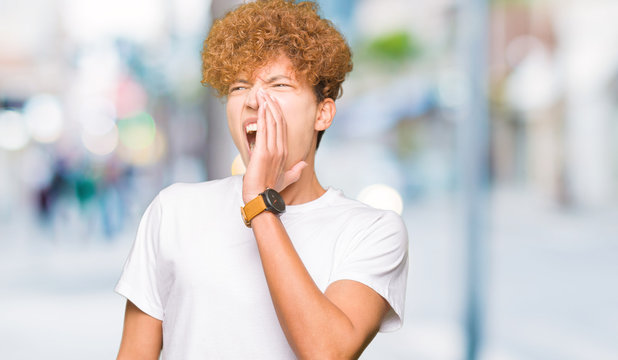 Young handsome man with afro hair wearing casual white t-shirt shouting and screaming loud to side with hand on mouth. Communication concept.