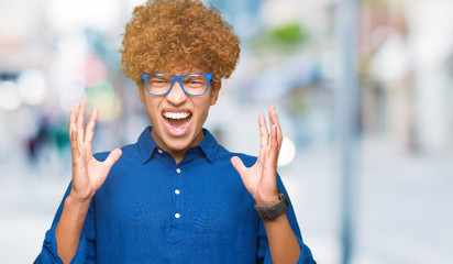 Young handsome man with afro hair wearing blue glasses crazy and mad shouting and yelling with...