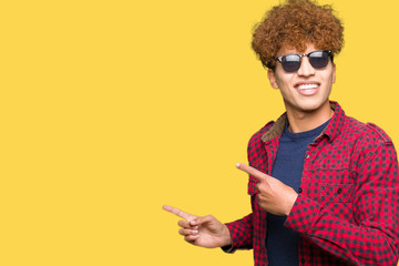 Young handsome man with afro hair wearing sunglasses smiling and looking at the camera pointing with two hands and fingers to the side.