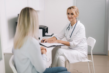 Young woman with blond hair at doctor appointment. Therapist female sitting at table and asks the patient questions