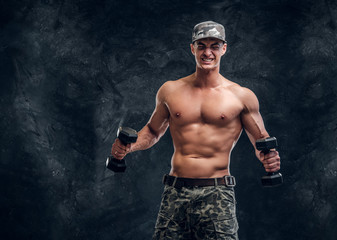 Attractive shirtless man in cap is doing exercises with dumbbells over dark background.