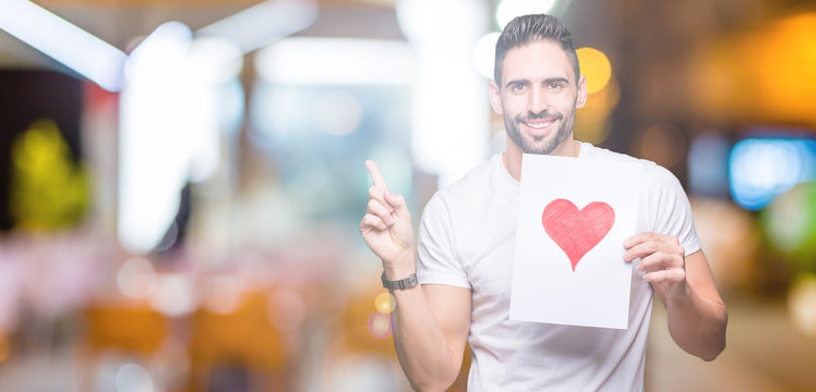 Handsome young man holding card with red heart over isolated background very happy pointing with hand and finger to the side