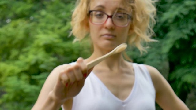 Young beautiful girl with ponytail is holding a useful bamboo toothbrush and looking at the camera outdoors during rainy weather on green tree background.