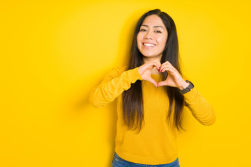 Beautiful brunette woman over yellow isolated background smiling in love showing heart symbol and shape with hands. Romantic concept.