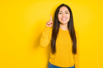 Beautiful brunette woman over yellow isolated background showing and pointing up with finger number one while smiling confident and happy.