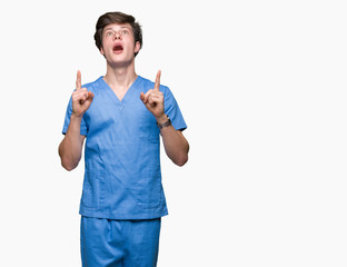 Young doctor wearing medical uniform over isolated background amazed and surprised looking up and pointing with fingers and raised arms.