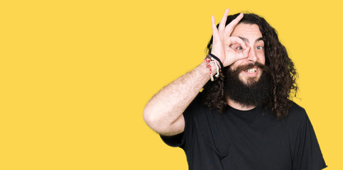 Obraz premium Young man with long hair and beard wearing heavy metal black outfit doing ok gesture with hand smiling, eye looking through fingers with happy face.
