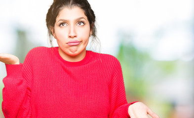 Young beautiful woman wearing red sweater and bun clueless and confused expression with arms and...