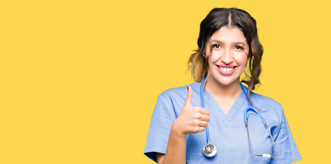 Young adult doctor woman wearing medical uniform doing happy thumbs up gesture with hand. Approving expression looking at the camera showing success.