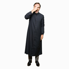 Middle age priest man wearing catholic robe peeking in shock covering face and eyes with hand,...
