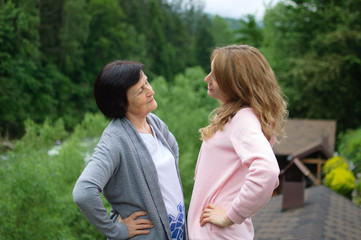 Outdoors portrait of happy mother and daughter looking to each other spending time together outside over landscape of forest and mountains.