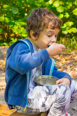 Cute white boy eats porridge from a bowl on a picnic outdoors