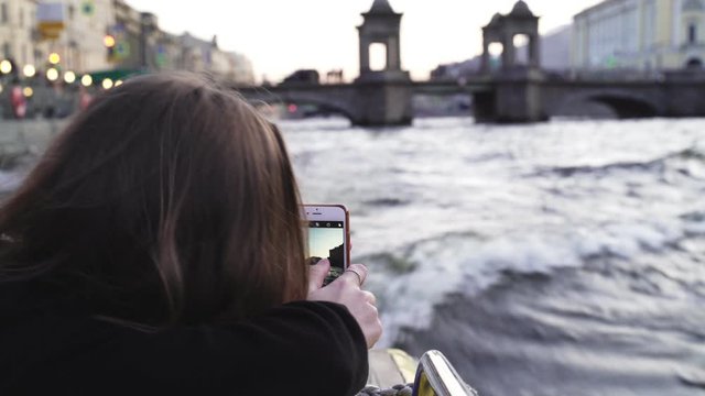 Girl making photos of water trip to rivers and channels of Saint Petersburg, Russia. Small waves and white foam from a ship engine. Parks, palaces and buildings on a background. Dark water, evening