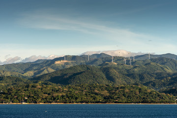 Caticlan, Malay, Philippines - March 4, 2019: Green mountain range with windmills dispersed under blue sky with thick gray cloudscape. Dark blue sea.
