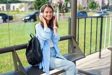 Teen girl listens to the music by white headphones in a public transport station while she waiting for tram.