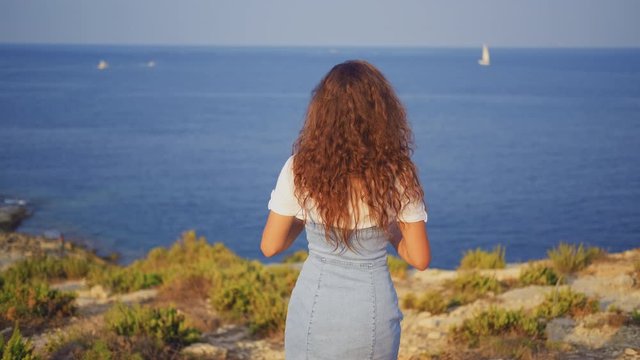 A young slender brunette woman stands on a cliff and photographs the sea and nature. Back view
