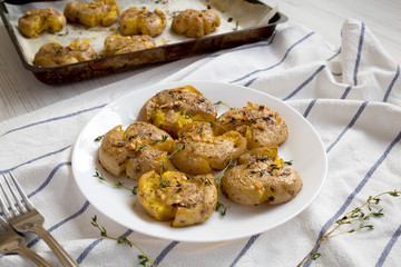 Homemade garlic thyme smashed potatoes on a white plate on a white wooden surface, side view. Closeup.