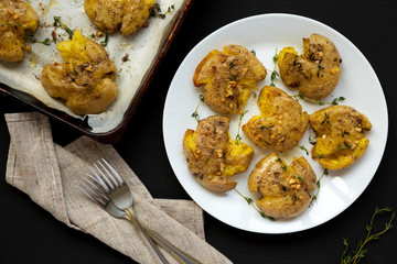 Homemade garlic thyme smashed potatoes on a white plate on a black surface, top view. Flat lay, overhead, from above. Closeup.