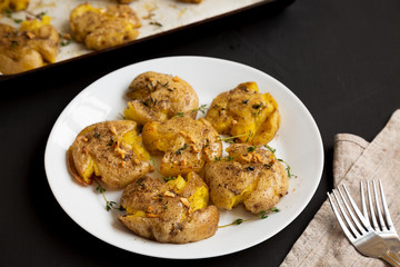 Homemade garlic thyme smashed potatoes on a white plate on a black surface, side view. Close-up.