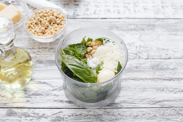 The process of making pesto sauce in a blender from cheese, pine nuts, olive oil, basil, garlic on the white wooden table.