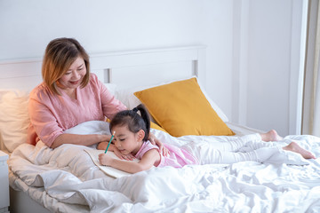 Selective focus happy little cute asian girl and her senior retired grandmother lying on the bed. Grandma smile and teach preschool grandchild do homework or write on the book on the bed in bedroom.