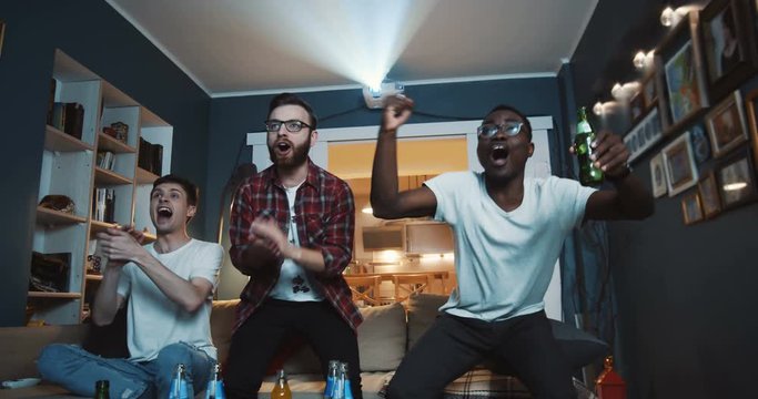 Happy young diverse male friends get crazy shouting for favourite sports team watching game on TV at home slow motion.
