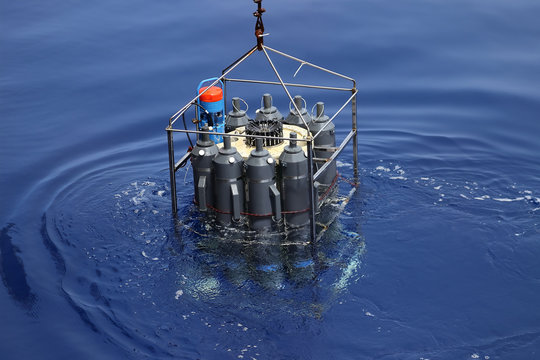 CTD rosette sampler with bathometers and other science equipment for measurement of underwater environment parameters like temperature, salinity and others. Sampler is half submerged in water.