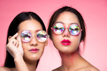 Two Korean Fashion Model looks at camera for shooting new collection Sunglasses.  Beautiful Asian 2 Women colorful make up wear Kaleidoscope Glasses with colorful, studio lighting pink background