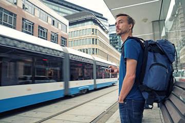 Young male traveller with backpack and smartphone stand on public transport stop and waiting tram in modern city center.