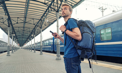 Fototapeta premium Young tourist backpacker with smartphone stand on railway station platform and waiting for train. Travel concept. Copy space.