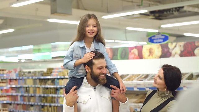 Young fashionable parents buy sweets at the store, the daughter sits on the shoulders of her father. Portrait of a happy family in a supermarket. Slow motion.