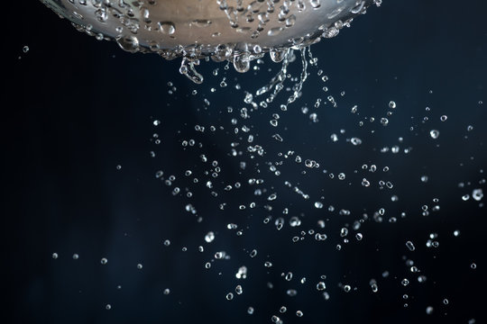 Flow and drops of water through holes in a colander on a dark background