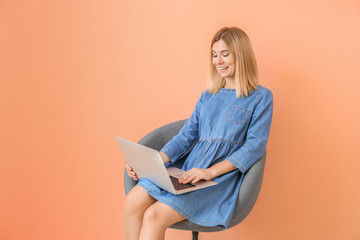 Happy woman with laptop sitting in armchair on color background