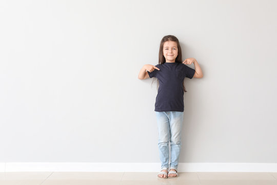 Cute little girl pointing at her t-shirt near light wall
