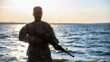 Silhouette of soldier in camouflage with assault rifle near river