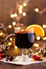 Composition with a glass of hot mulled red wine with spices. Holiday atmosphere, christmas ligths on dark background. Rustic style, cones, nuts, anise, cinnamon, orange as festive decor 
