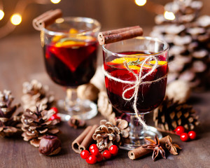 Composition with two glasses of hot mulled red wine with spices. Holiday atmosphere, christmas ligths on dark background. Rustic style, cones, nuts, anise, cinnamon, orang, berries as festive decor 