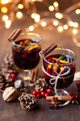 Composition with two glasses of hot mulled red wine with spices. Holiday atmosphere, christmas ligths on dark background. Rustic style, cones, nuts, anise, cinnamon, orang, berries as festive decor 