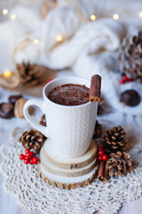 Composition with white mug with hot chocolate with spices. Decor rustic style with natural elements: cones, red berries, wood, cinnamon sticks, nuts. Christmas lights on, knitted plaid, cozy morning