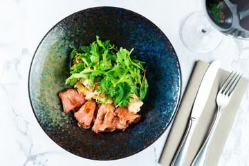 Warm salad with roast beef, crackers, arugula and sauce in a black bowl on a marble table served with red wine. Tasty and healthy snack for lunch. Food photo background