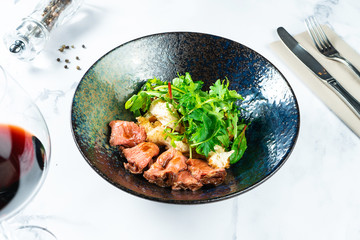 Warm salad with roast beef, crackers, arugula and sauce in a black bowl on a marble table served with red wine. Tasty and healthy snack for lunch. Food photo background