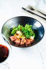 Warm salad with roast beef, crackers, arugula and sauce in a black bowl on a marble table served with red wine. Tasty and healthy snack for lunch. Food photo background